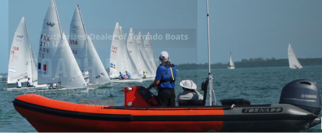 A person in a blue shirt steers an orange and black inflatable motorboat labeled 'TORNADO' near a group of sailboats racing on the water. The sky is clear and trees are visible in the distance.