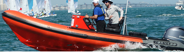 Two men ride a bright orange inflatable motorboat named “Tornado” on choppy blue water, with several sailboats and a city skyline visible in the distant background.