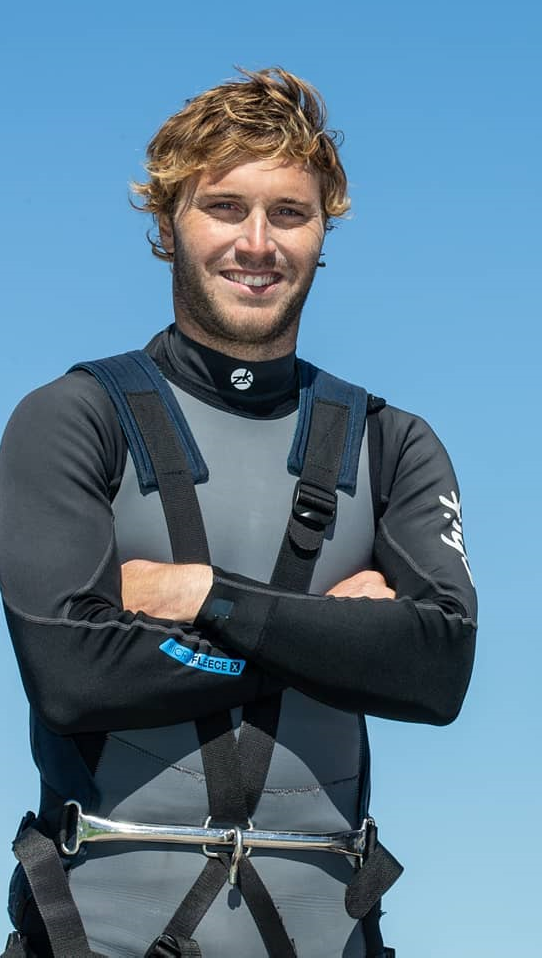 A man with wavy blond hair wearing a black and gray wetsuit and harness stands outdoors with his arms crossed, smiling, against a clear blue sky.