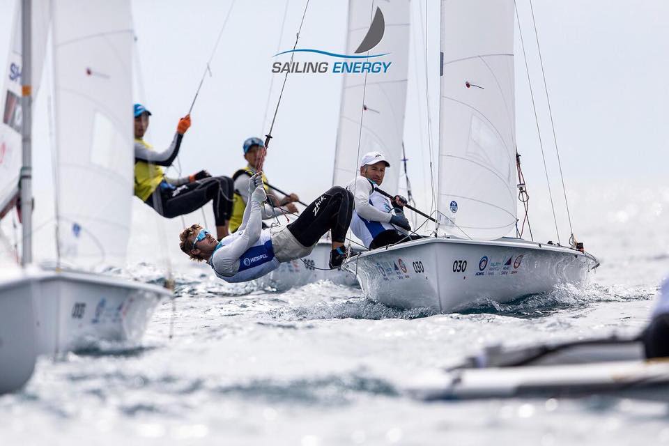 Several teams compete in a sailing race, maneuvering white sailboats on the water. Sailors lean out over the edge of their boats, holding ropes, with sails billowing and 'SAILING ENERGY' logo visible above.