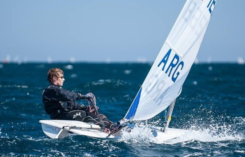 A sailor steers a small white sailboat with “ARG” on the sail across choppy blue water, wearing sunglasses and a black wetsuit, with a clear blue sky in the background.