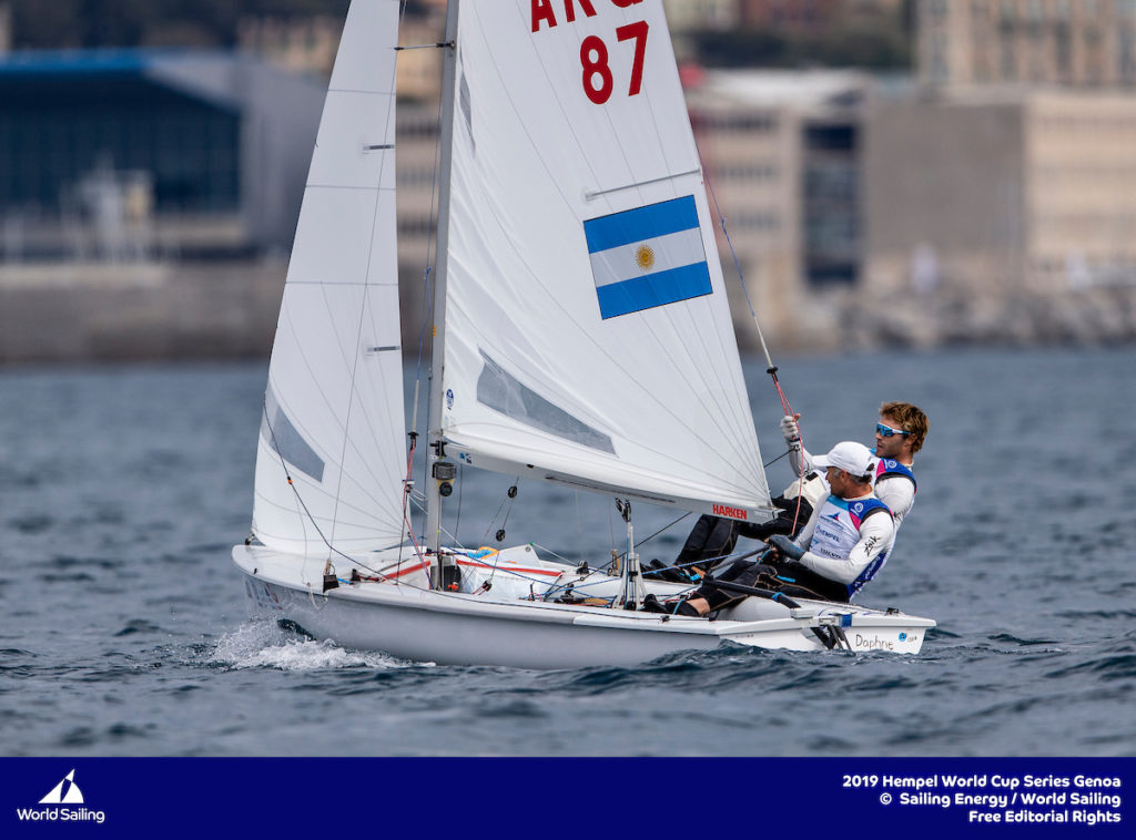 Two sailors compete in a small sailboat displaying the Argentine flag, leaning out over the water during a race at the 2019 Hempel World Cup Series in Genoa. Buildings and hills are visible in the background.