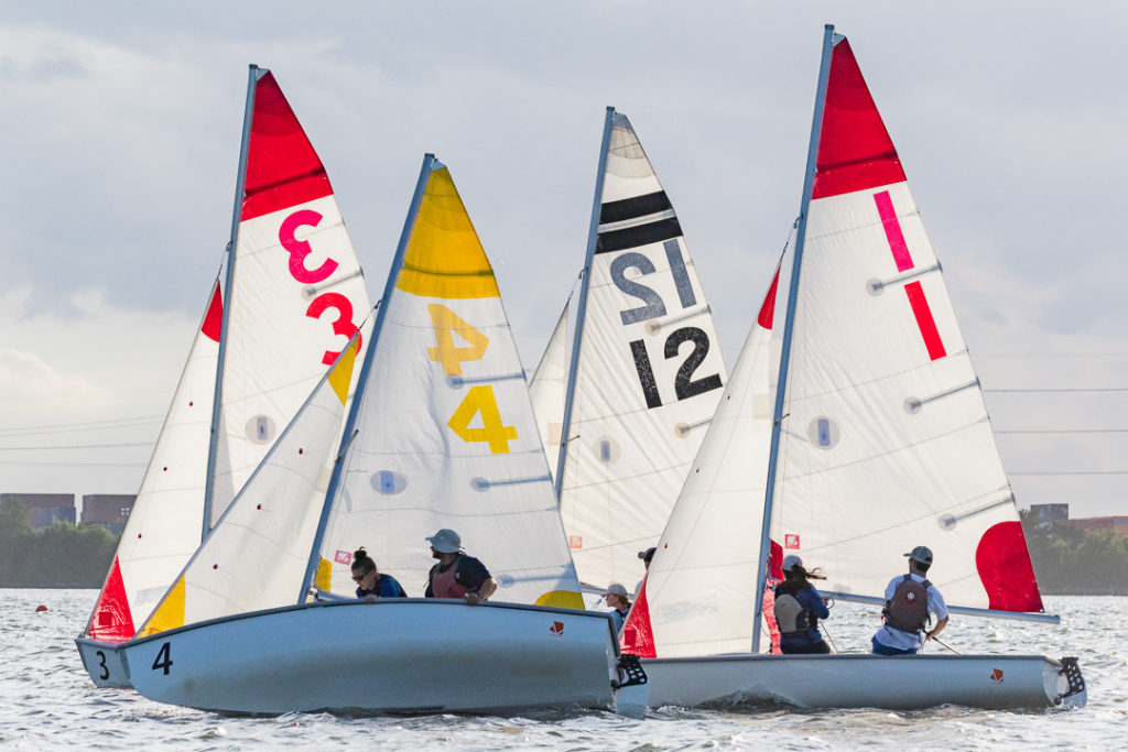 Four small sailboats with colorful numbered sails (3, 4, 12, 1) are racing closely together on a body of water. Each boat has two sailors, and the background shows a cloudy sky and distant shoreline.