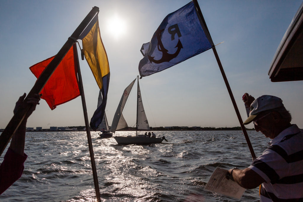 Two people on a boat hold colorful flags with sailboats racing in the background under a bright sun in a slightly cloudy sky, with choppy water and a distant shoreline visible.