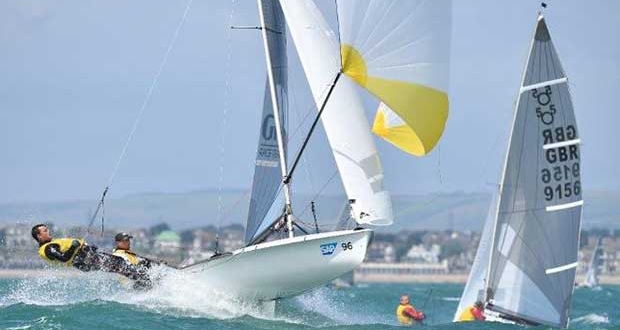 Two sailboats race on a sunny, windy day with sailors leaning out over the side for balance. A town and hills are visible in the background, and another smaller boat is in the water nearby.
