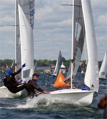 Two people sail a small racing sailboat around an orange buoy on a lake, leaning out over the water as other sailboats compete nearby. The sky is partly cloudy and trees line the distant shore.