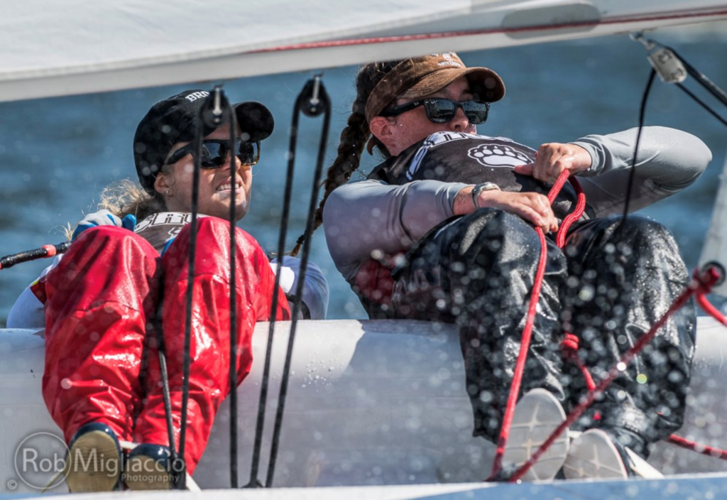 Two sailors in sunglasses and hats lean back in a small sailboat, wearing red waterproof pants, with water splashing on the lens and blue water in the background.