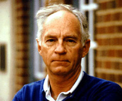 An older man with short gray hair wearing a blue collared shirt stands outdoors in front of a brick building, looking directly at the camera with a neutral expression.