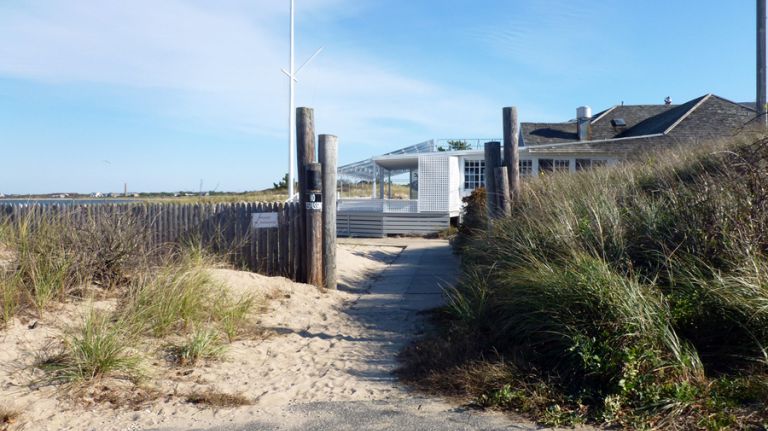 A sandy path lined with tall grass and wooden posts leads to a white building by the beach, with a blue sky overhead and a glimpse of water on the left.