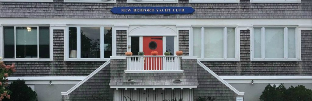 The front entrance of the New Bedford Yacht Club, featuring a red door, a small balcony with pumpkins on the railing, large windows, and a blue sign with the club's name above the entrance.