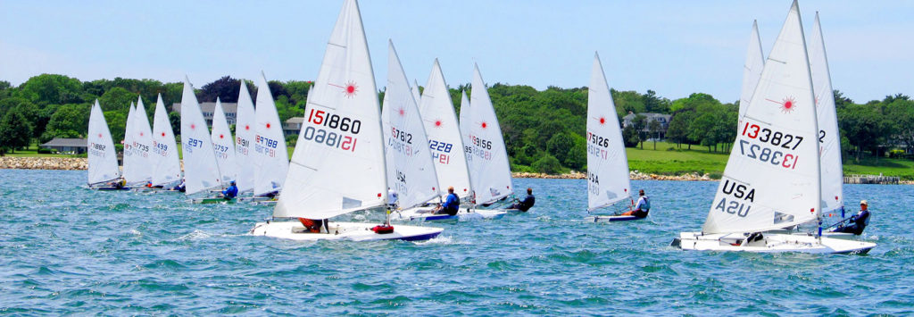 A group of small sailboats with white sails race on a blue lake near a green, tree-lined shore under a clear sky. Several sailors are visible in each boat.