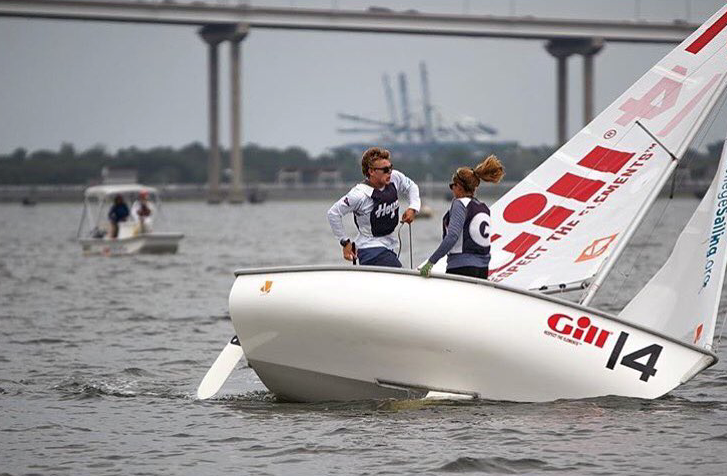 Two sailors in matching uniforms navigate a small sailboat labeled “Gill 14” on the water near a bridge, with another boat and industrial cranes visible in the background.