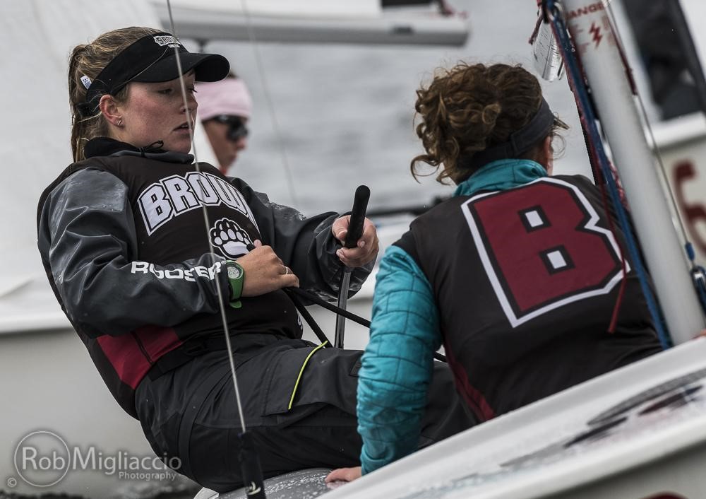 Two female sailors wearing 'Brown' team vests compete in a sailing race, focusing intently as they handle ropes and steering equipment on a sailboat. One faces forward, the other’s back shows a large “B” on her vest.