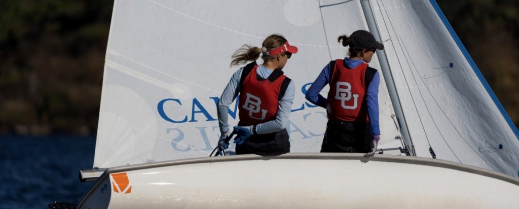 Two people wearing red BU vests and caps are sailing a small white sailboat on a lake. They are looking in opposite directions, and the boat’s sail has blue text partially visible.