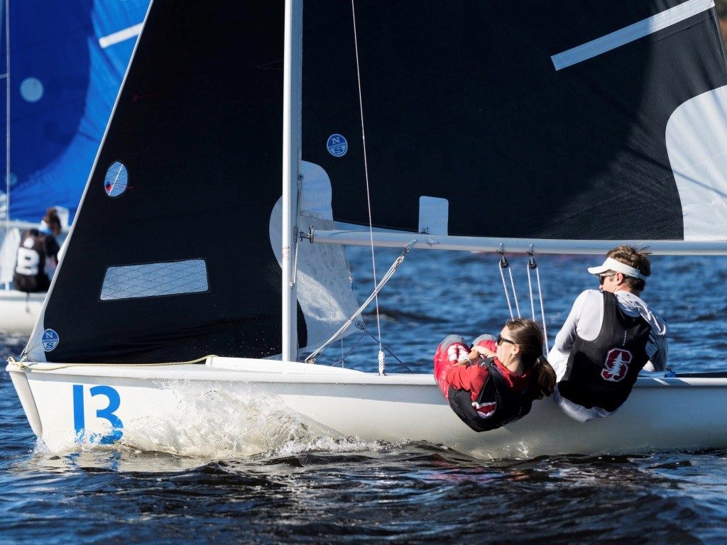 Two sailors in life vests and sunglasses lean out of a small sailboat numbered 13, gliding through the water with black sails during a sunny day, as other boats race in the background.