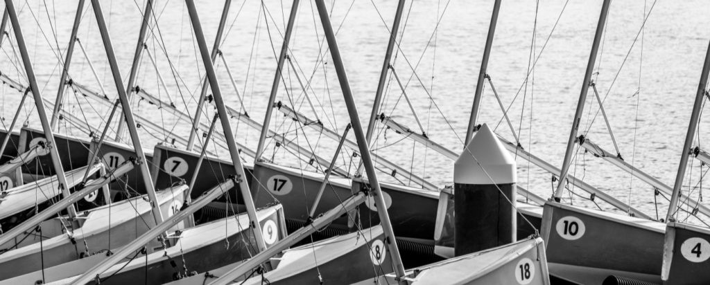 Black and white photo of small sailboats docked in a row, each with a visible number on the hull. Sail masts are upright and closely spaced, with calm water in the background.