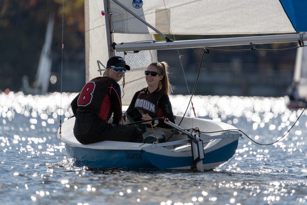 Two people in sailing gear are smiling as they navigate a small sailboat named "Joanne" on a sunlit, sparkling body of water, with other boats and blurred trees in the background.