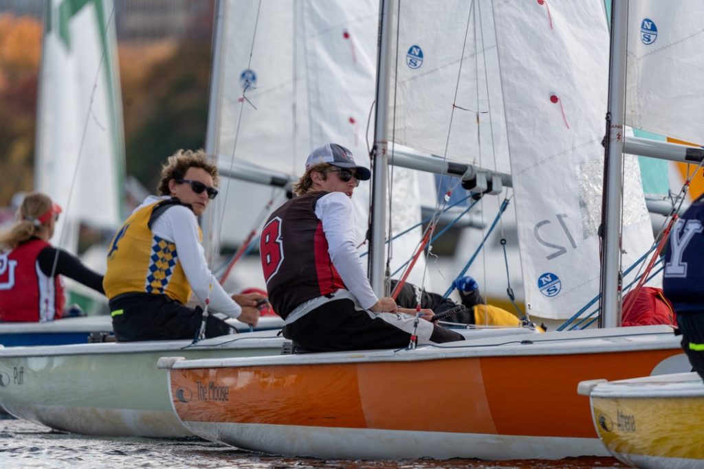 Several sailors wearing team vests and sunglasses compete in a sailboat race on a sunny day, focused and adjusting their positions in their boats as they navigate close together on the water.