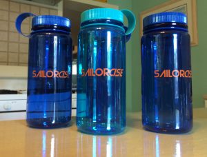 Three blue water bottles with orange 'SAILORGISE' text, standing on a light-colored countertop in a kitchen setting. The bottle on the left has a blue lid, the middle one has a turquoise lid, and the right one has a blue lid.