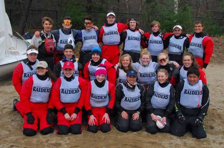 A group of 20 young people in red and black sailing gear and gray "Raiders" vests pose together outdoors on a sandy area, smiling at the camera. Trees and a sailboat are visible in the background.