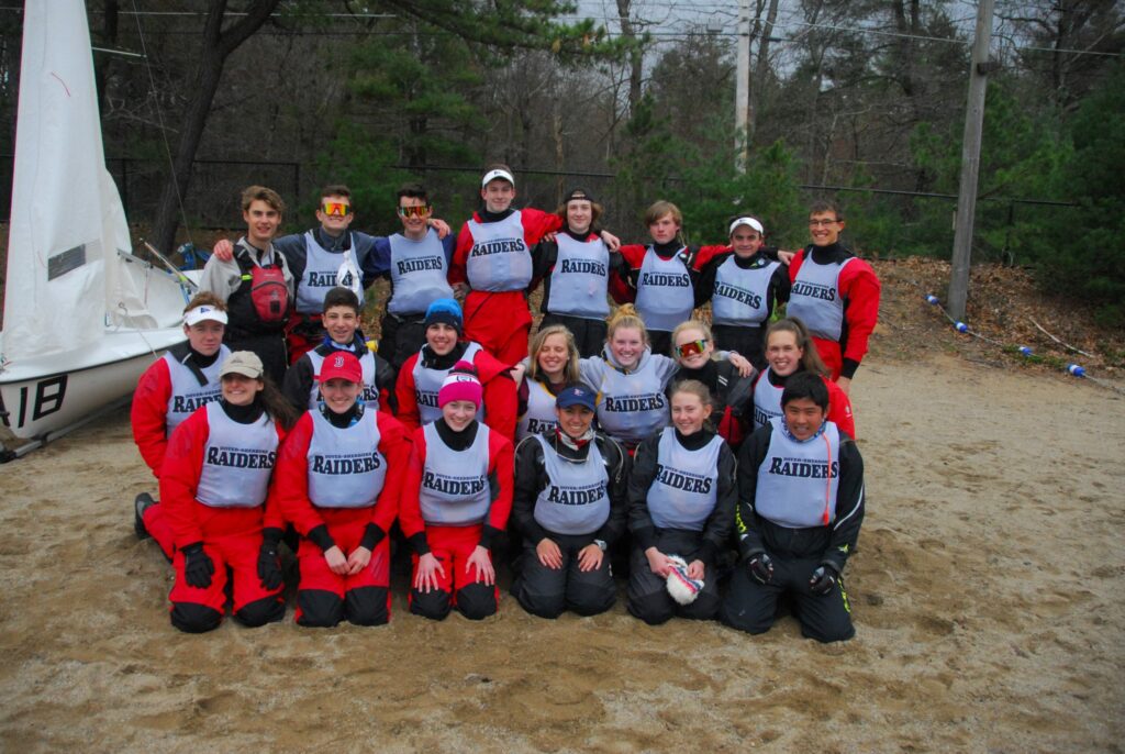 A group of young sailors wearing red and black gear and “RAIDERS” vests pose outdoors on sandy ground with a sailboat and trees in the background.