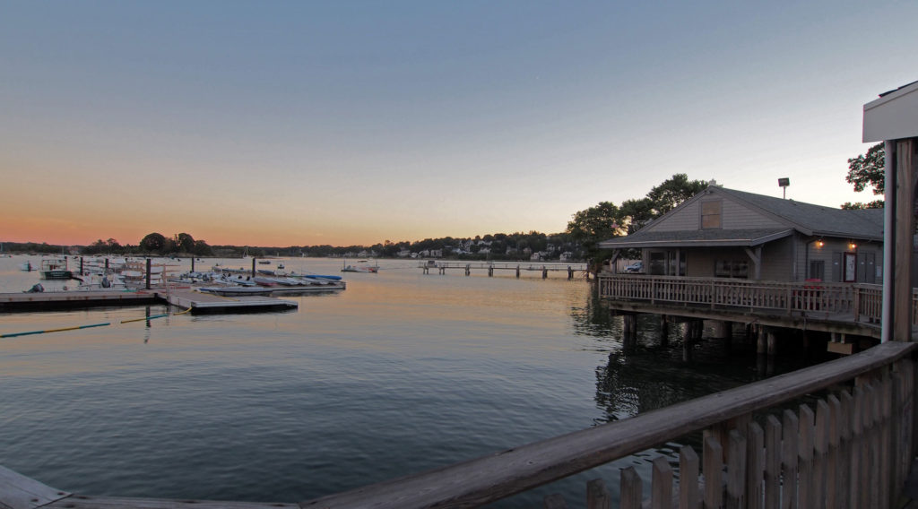 A calm waterfront scene at sunset with a wooden building on stilts over the water, several docks with boats, and trees lining the opposite shore under a clear sky.