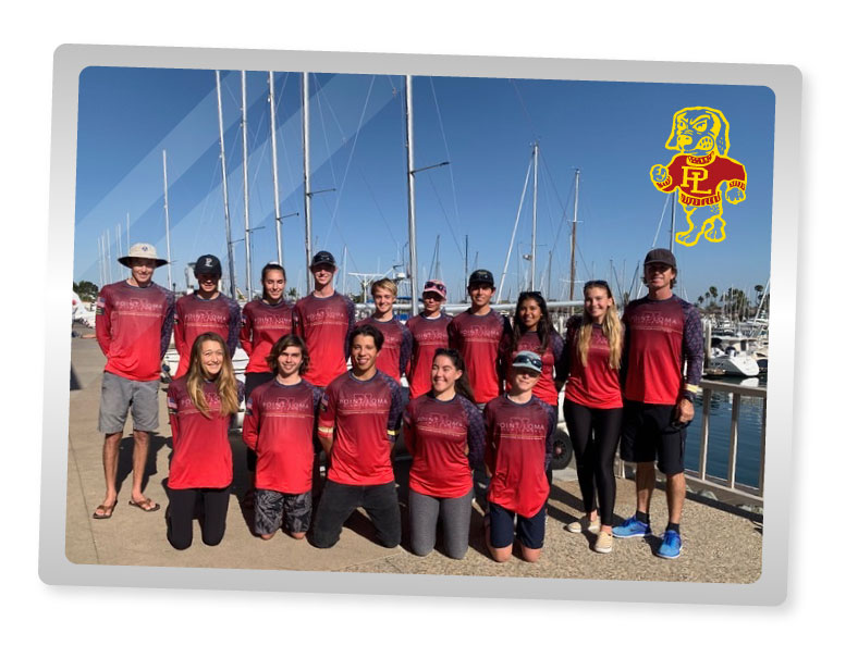 A group of thirteen people in matching red shirts poses outdoors by a marina with sailboats. They are standing and kneeling on a sidewalk, with a lion mascot logo in the top right corner.
