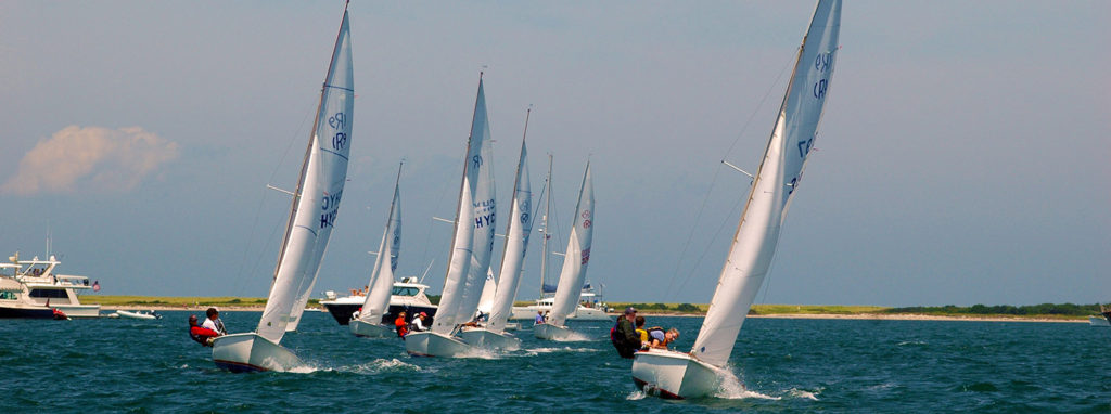 Several sailboats with people onboard race closely together on blue water, with other boats and a low, grassy shoreline visible in the background under a partly cloudy sky.