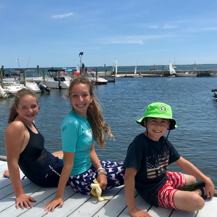 Three smiling kids sit on a dock by the water, with boats and sailboats in the background under a blue sky. The boy wears a green hat; the two girls wear swimsuits and look at the camera.
