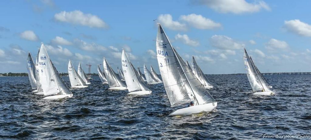 A group of small sailboats with white sails racing on a blue, slightly choppy body of water under a partly cloudy sky. Land and a bridge are visible in the distant background.