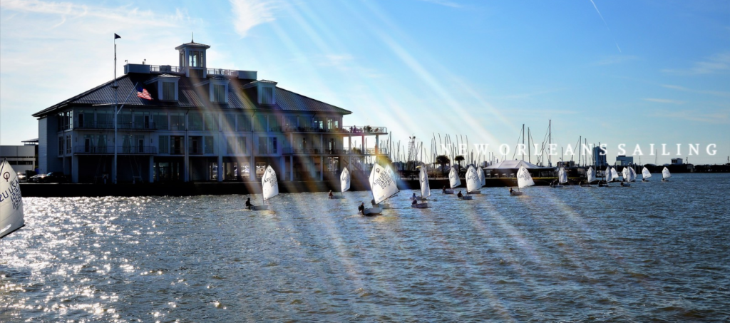 Sailboats race on a sunlit lake near a large building with an American flag, with text “NEW ORLEANS SAILING” in the background and light rays shining over the scene.
