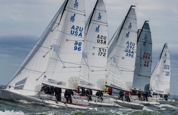 Several sailboats with USA markings race closely together on choppy water under a cloudy sky. Each boat has a crew working at the sails, and the boats display large white sails with numbers.