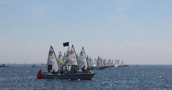 A fleet of small sailboats with white sails prepares to race on calm blue water, following a larger boat with a blue flag. The sky is mostly clear, and more boats are visible in the distance.