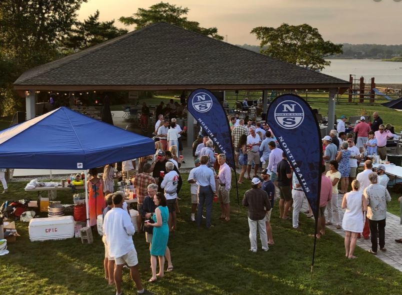 A large group of people gather outdoors under tents and a pavilion near the water at sunset. Some attendees are socializing, others stand by tables with food and drinks. Two blue flags with an "N" and "NORTHEASTERN" are visible.