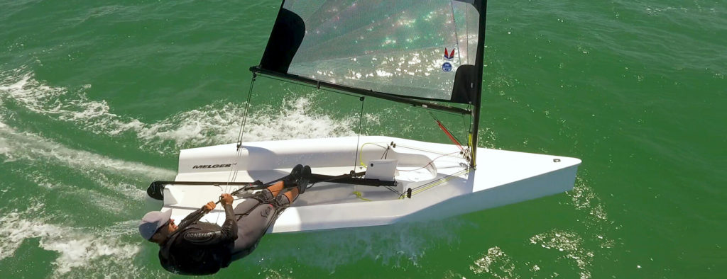 A person sails a small, white racing dinghy over green water, leaning out over the edge to balance the boat. The dinghy has a transparent sail and is moving quickly, creating ripples and foam behind it.