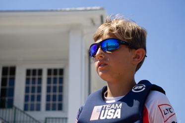 A young boy wearing reflective sunglasses and a Team USA life vest stands outside in sunlight, with a white building and blue sky in the background.