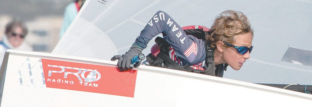 A sailor wearing sunglasses and a "TEAM USA" jersey leans out of a racing sailboat, gripping the edge and looking ahead with focus during a competition.