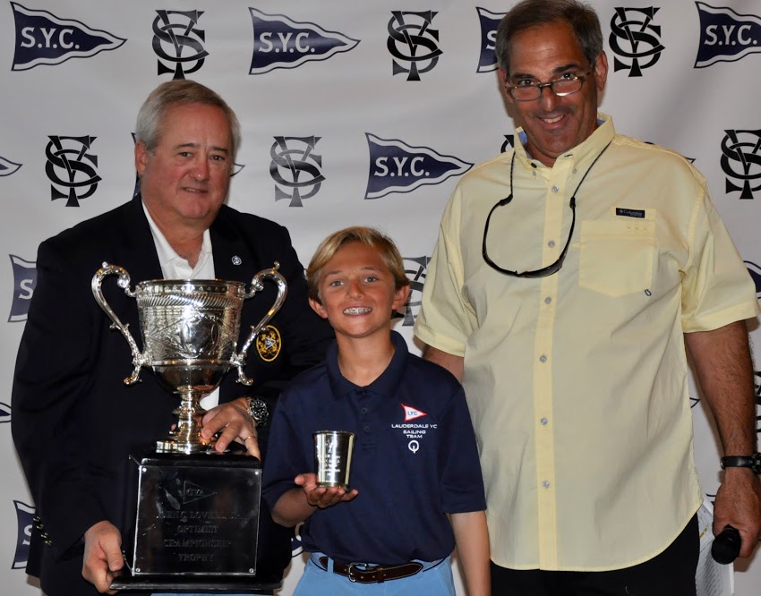 Three people stand smiling in front of an SYC-branded backdrop. The man on the left holds a large silver trophy, the boy in the middle holds a smaller cup, and the man on the right stands beside them wearing glasses and a yellow shirt.