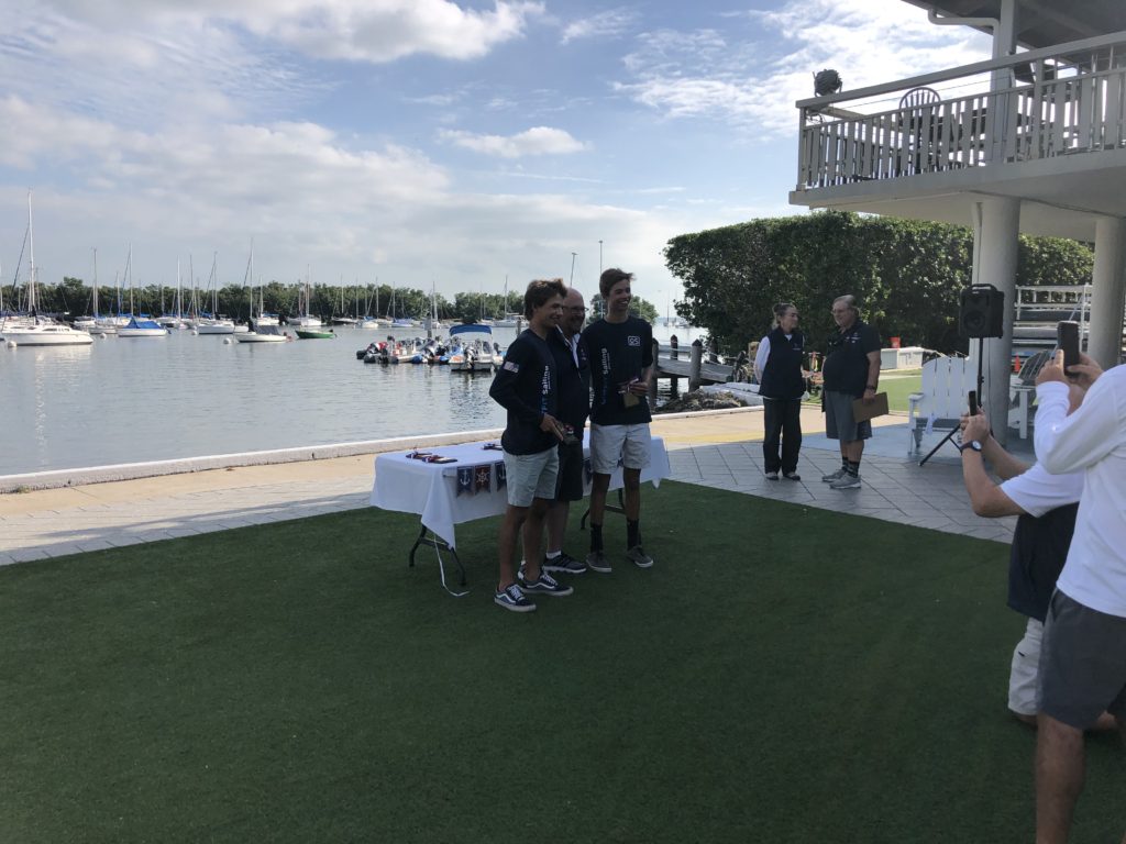 Three people pose for a photo near a marina with sailboats, standing on grass by a table with awards. Other people watch, and the scene is sunny with partly cloudy skies.