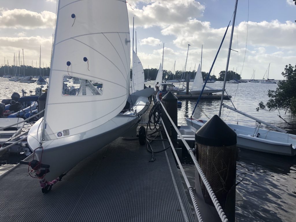 Several sailboats are docked at a marina, with their white sails raised. The boats are tied to a floating metal dock surrounded by water, and trees and many other sailboats are visible in the background under a partly cloudy sky.
