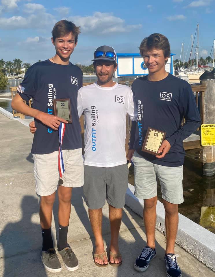 Three people stand smiling at a marina. Two teens in navy “Outfit Sailing” shirts each hold a plaque; an adult in a white shirt stands between them. Boats and palm trees appear in the background under a blue sky.