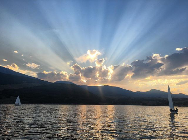 Two sailboats glide on a calm lake at sunset, with dramatic sun rays streaming through clouds above distant mountains, creating a striking and serene scene.