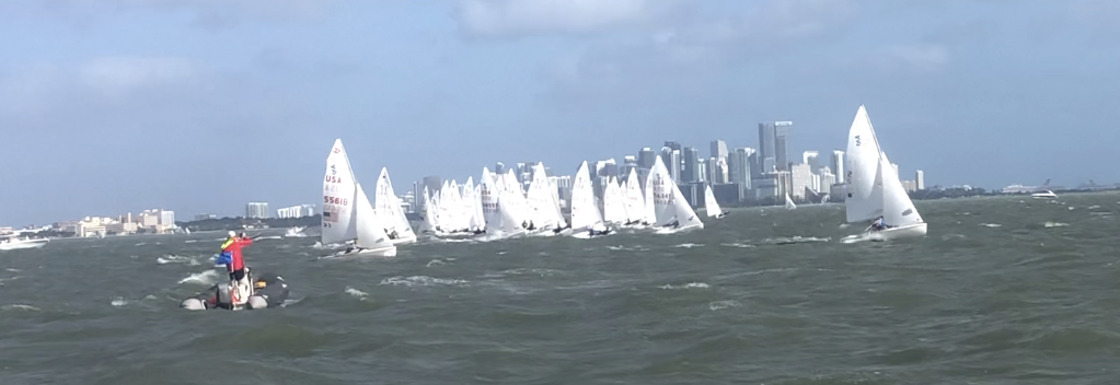 A group of sailboats races on choppy water near a city skyline under a partly cloudy sky. A small motorboat with people in bright vests watches from the left. The city buildings rise in the background.