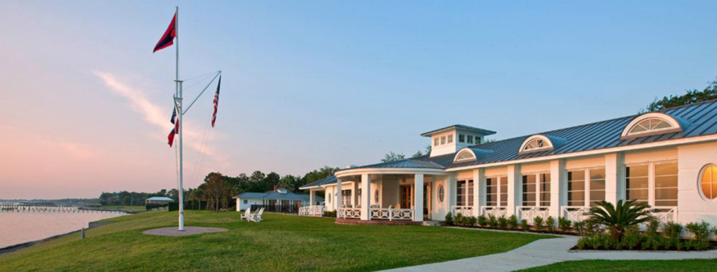 A waterfront clubhouse with white walls and a metal roof sits beside a calm shoreline at sunset, with flags on tall poles and Adirondack chairs on the lawn. Trees line the background.