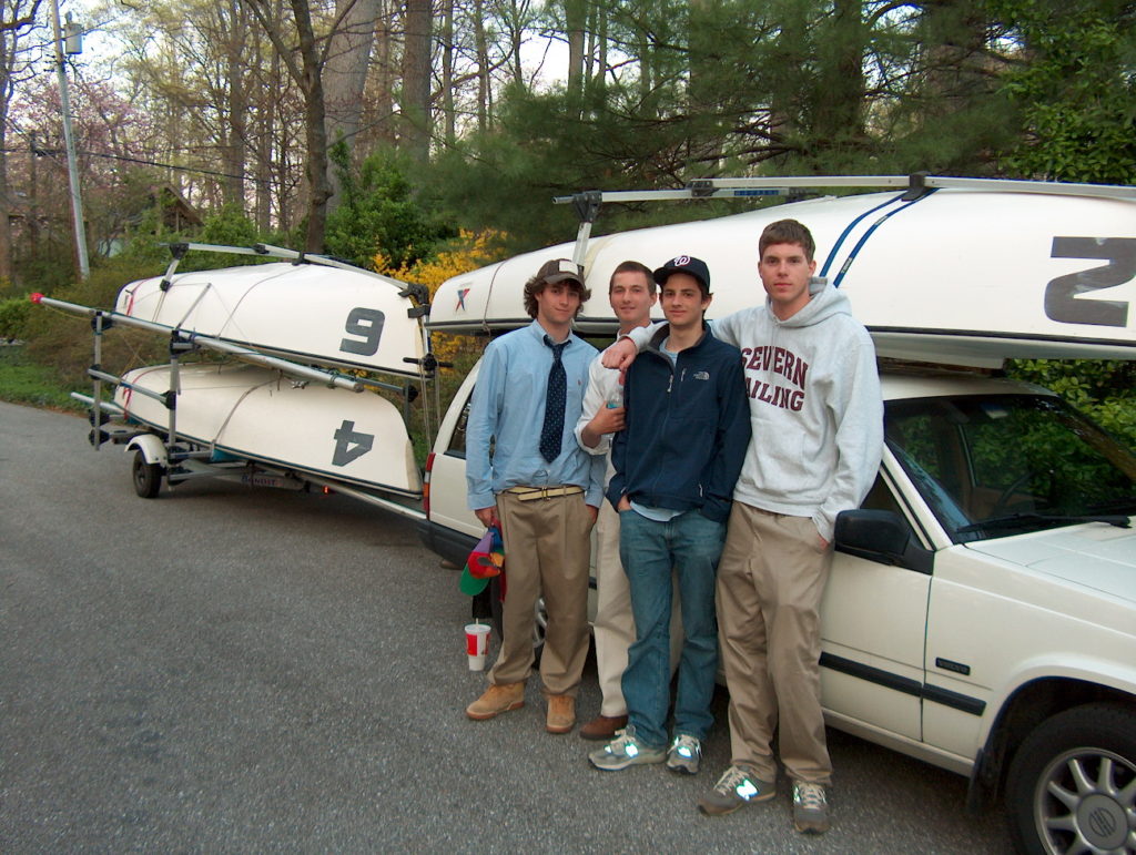 Four young men stand together beside a white car towing a trailer with small sailboats stacked on it. They are outdoors on a paved road surrounded by trees. Some are dressed casually; one wears a tie.