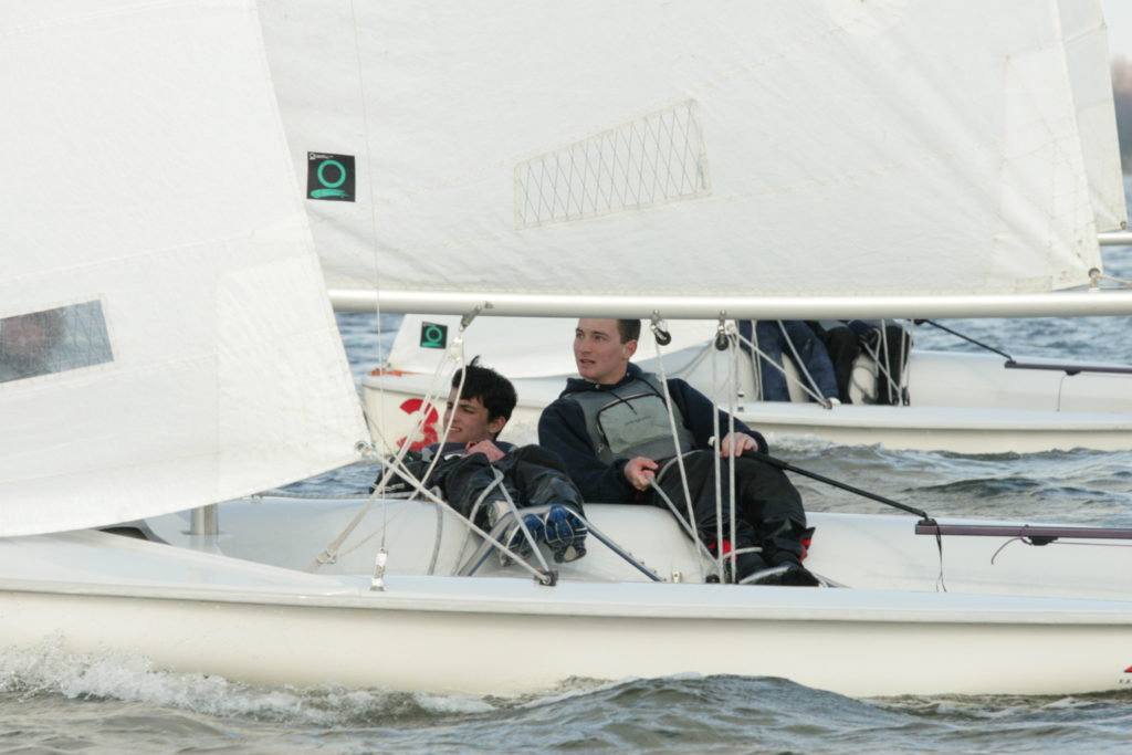 Two young sailors sit in a small sailboat, steering and adjusting the sails while racing on choppy water. Another sailboat with two people is visible in the background. Both boats have white sails.
