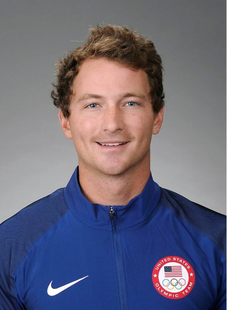 A smiling man with short, curly brown hair wears a blue Nike jacket with a 'United States Olympic Team' logo, posing in front of a neutral gray background.