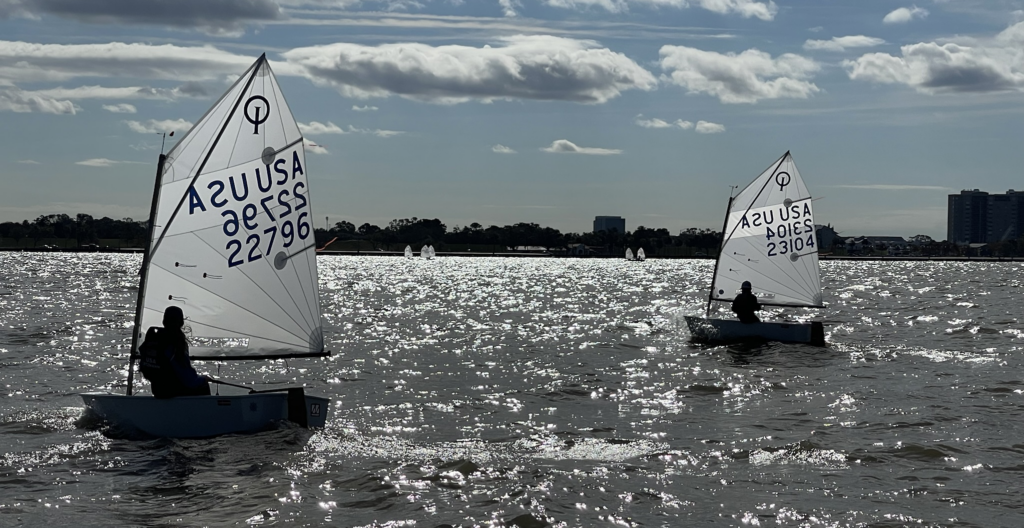 Two small sailboats with young sailors on a sparkling lake, sails marked 'USA' and sail numbers visible. The sky is partly cloudy and buildings are seen in the distant background.