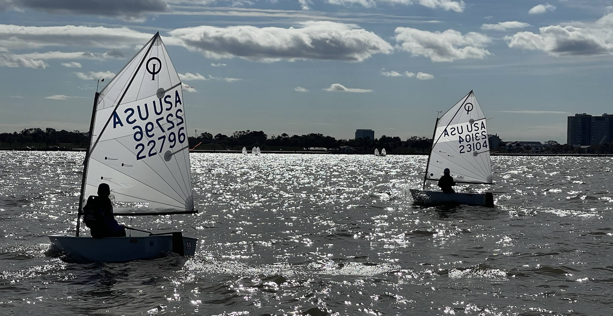 Two small sailboats with young sailors on a sparkling lake, sails marked "USA" and sail numbers visible. The sky is partly cloudy and buildings are seen in the distant background.