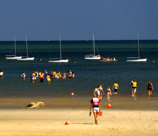 People wearing life jackets walk toward the sea from a sandy beach, while groups gather in the shallow water. Several sailboats float in the background under a clear sky.
